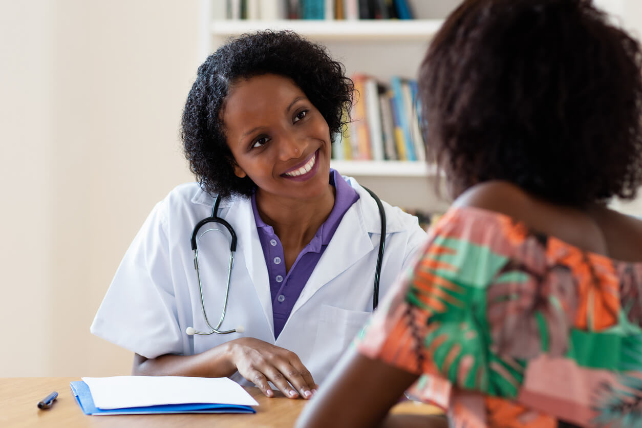 A black woman gynecologist speaks with a black woman patient about scheduling her pap smear. The gynecologist is facing towards the viewer, and can be seen smiling, while the patient is facing away.