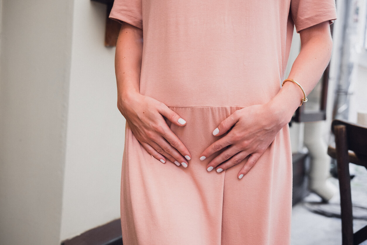 A woman holding her abdomen walks towards an exam room for a pelvic exam. The woman is wearing a pink outfit.