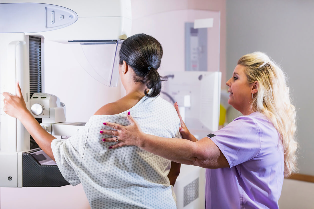 A woman prepares for a mamogram. She is wearing a hospital gown and is being assisted by a woman nurse wearing purple scrubs